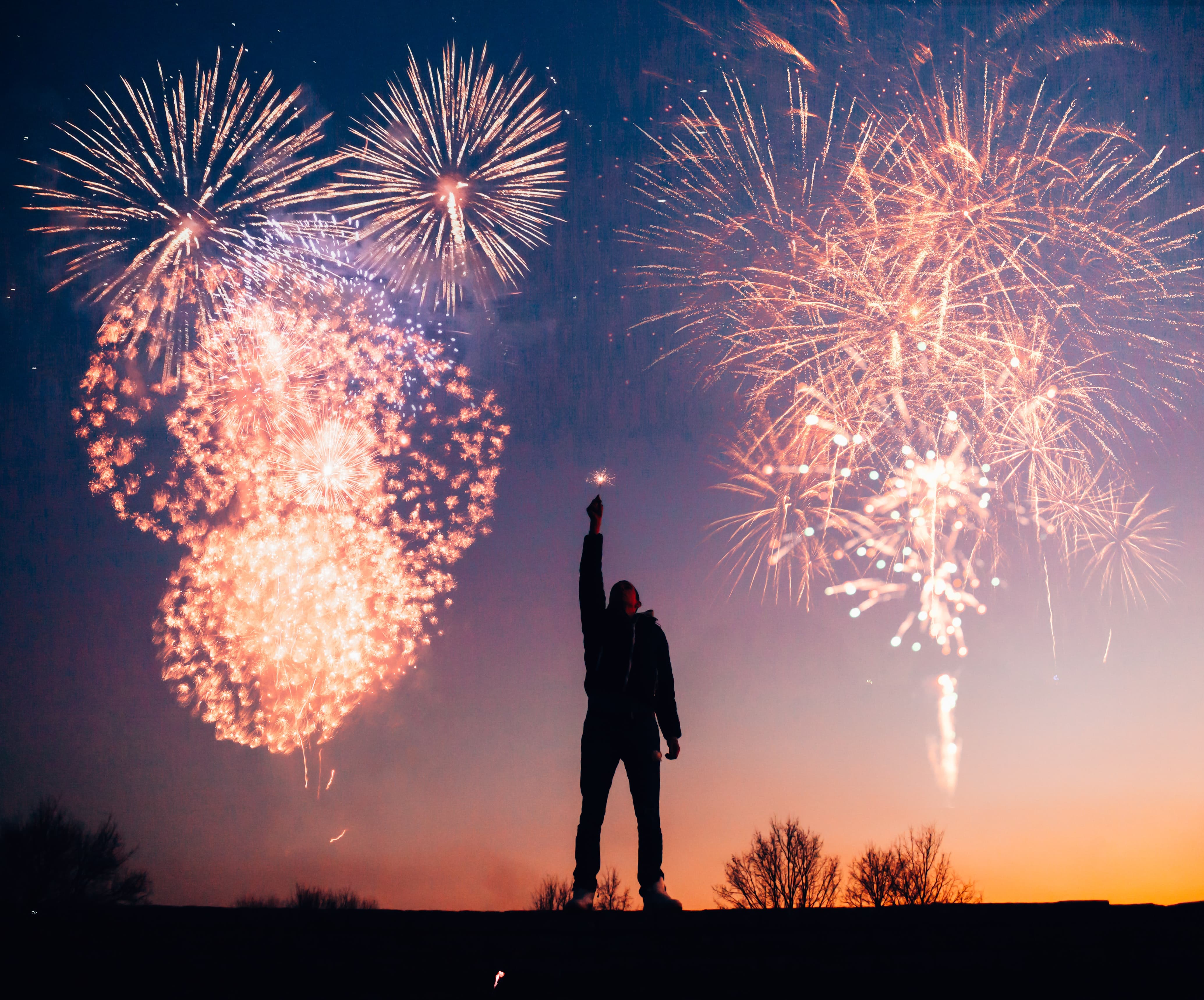 Multiple fireworks in the sky with a man holding a sparkler on top of a hill at twilight